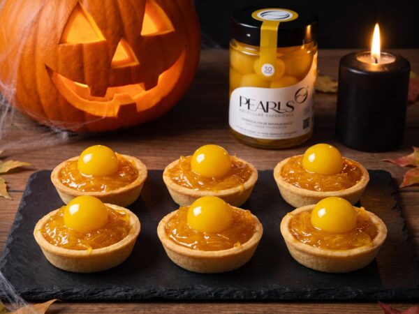 Seis tartaletas de confitura de calabaza con una Pearl de Naranja Dulce encima, sobre pizarra, junto al frasco y una calabaza de Halloween